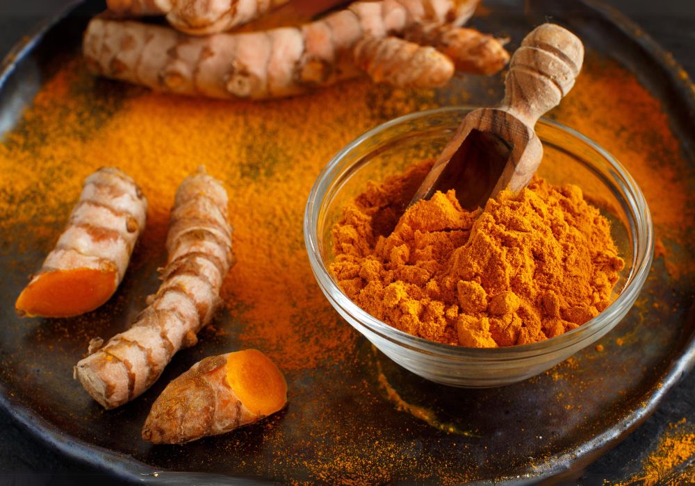 Em uma bandeja, um pote de vidro com cúrcuma em pó e raízes de cúrcuma ao lado, dentro da bandeja.