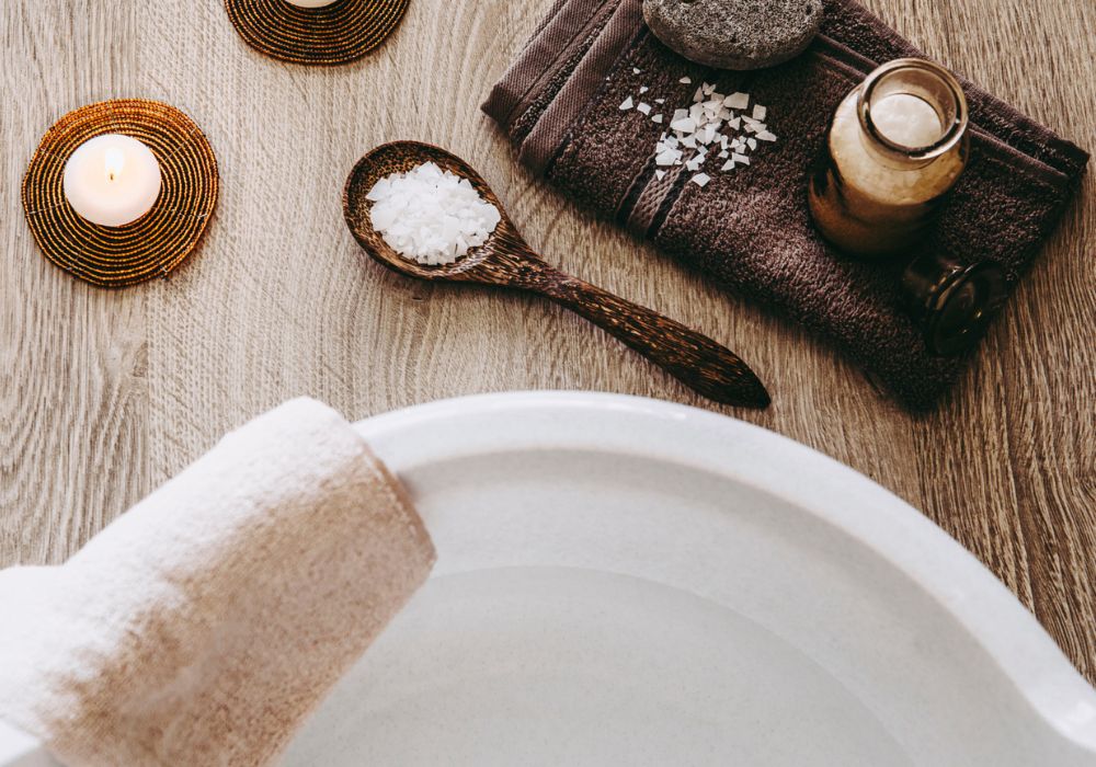 Bacia um uma toalha pendurada e ao lado um pote com sal grosso, uma colher de madeira com sal grosso e uma vela aromática acessa.