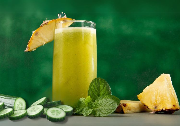 Um copo de suco com abacaxi, hortelã e pepino/ E todos os ingredientes ao redor. Todos em cima de uma mesa.