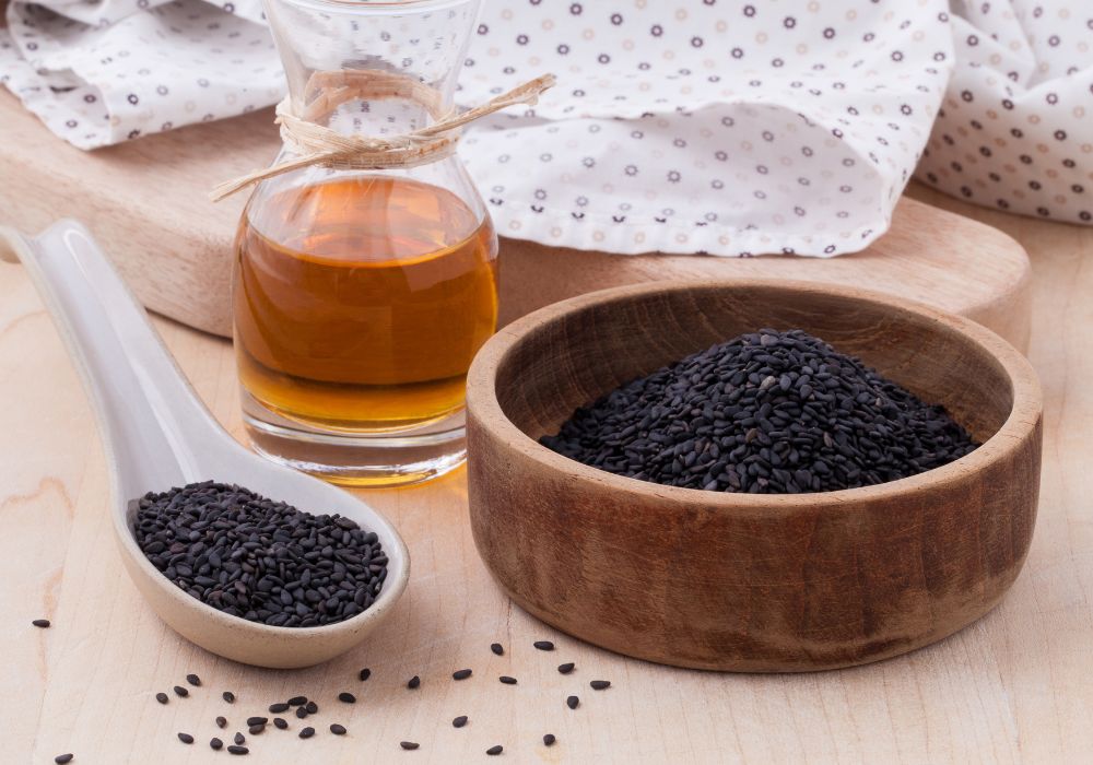 Um vidro com Óleo de Gergelim; um pote com Gergelim preto e uma colher de plástico com um punhado de gergelim, também preto.