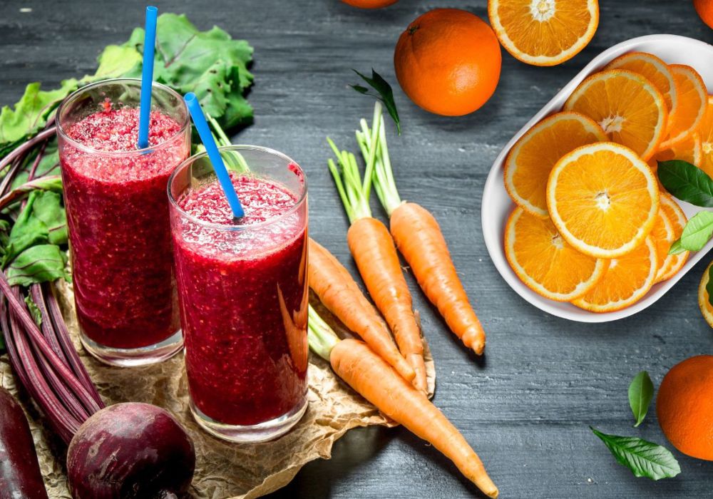 Dois copos de sucos de beterraba, laranja e cenoura com os ingredientes ao redor.