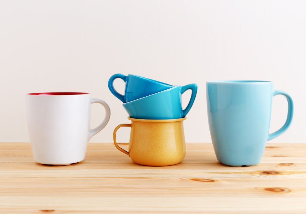 Xícaras e canecas. Da esquerda para a direita uma caneca branca com a parte interna na cor vermelha; no meio três xícaras uma em cima da outra. Duas nas cores azuis e uma na cor amarela; e, por último, uma caneca na cor azul clara.