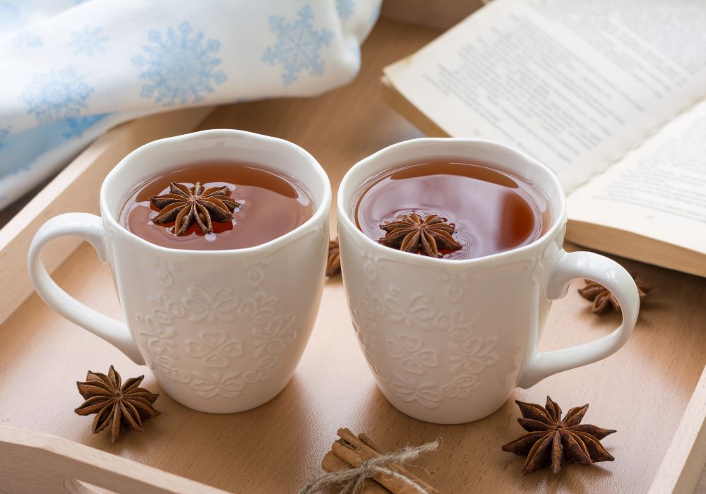 Duas canecas brancas com chá de Anis Estrelado em uma bandeja; ao lado alguns anis estrelados e ao fundo um livro aberto.
