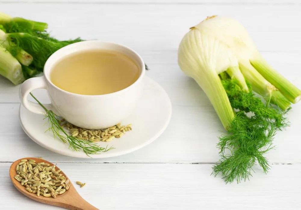 Uma xícara branca de porcelana com chá de erva-doce dentro , uma colher de madeira com ervas-doce dentro e uma raiz da planta de erva-doce ao lado.