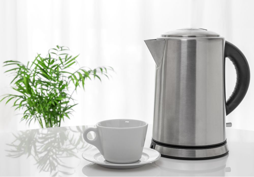 Uma chaleira elétrica prata e uma xícara branca de porcelana ao lado.