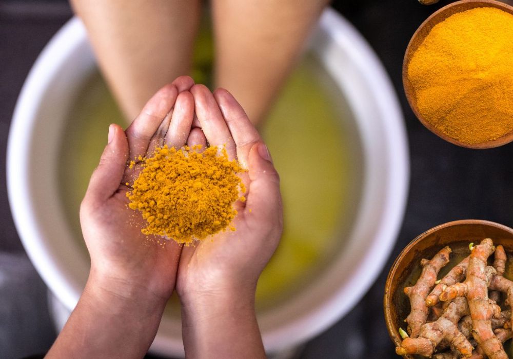 Uma pessoa com os pés dentro de uma bacia com água e uma pessoa segurando o pó do açafrão em nas mãos, e no chão dois potes, sendo um com açafrão em pó e outro com açafrão em raiz.