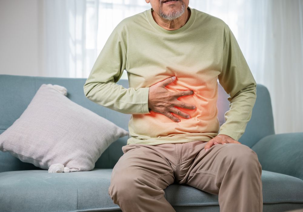 Um homem sentado no sofá com a mão direita na altura do estômago, indicando dor. Gastrite.