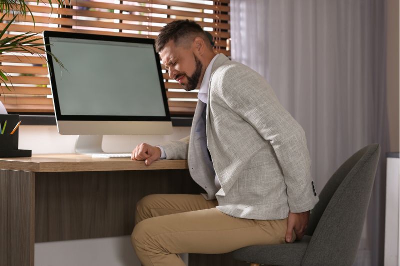 Homem sentado em mesa de escritório, demonstrando dor por estar com hemorroida.