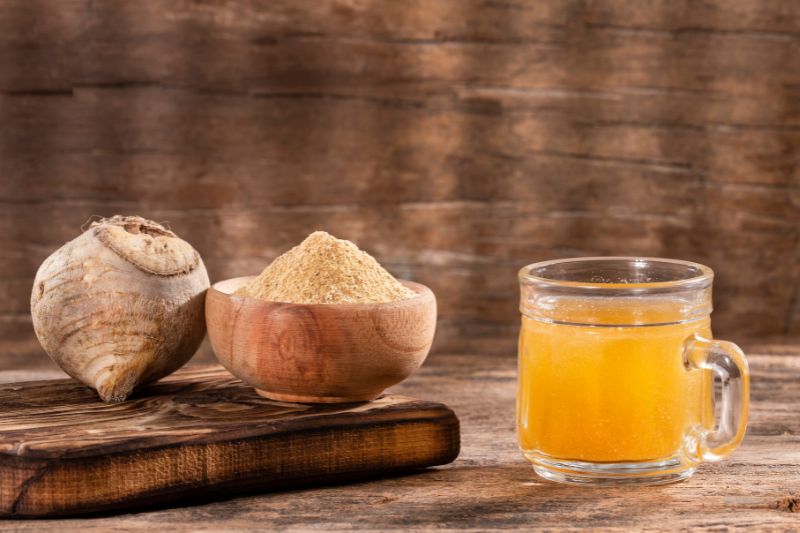 Uma tábua de madeira com o tubérculo da maca peruana em cima e um pote pequeno de madeira com maca peruana em pó e um copo ao lado com suco que consta maca peruana em pó.